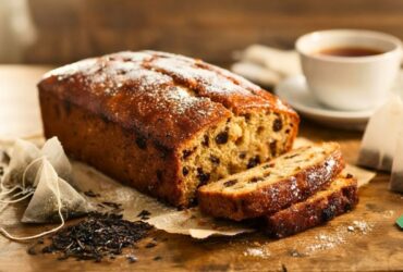 Golden-brown Mary Berry Earl Grey Tea Loaf Recipe Loaf on a wooden table, with one slice cut to show soft, moist interior. Light dusting of icing sugar, Earl Grey tea bags scattered nearby, and a blurred cup of tea in the background, warm natural lighting, cozy kitchen setting.