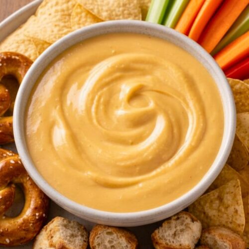 Warm bowl of creamy golden beer cheese dip with steam rising, surrounded by soft pretzels, bread pieces, and chips on a rustic wooden table.